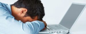 A man in a blue shirt rests his head on his folded arms in front of an open laptop, appearing tired or frustrated—possibly overwhelmed by the things people hate about managing SEO on a website.