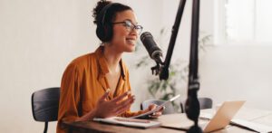 A woman wearing glasses and headphones speaks into a microphone in a recording studio, perhaps creating her church podcast. She sits at a table with a laptop, notebook, and pen in the bright, modern setting with soft lighting and a blurred background.
