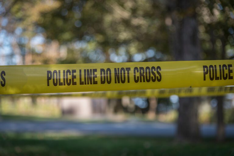 Yellow police tape with "POLICE LINE DO NOT CROSS" blocks a wooded area, reinforcing the importance of church safety in outdoor settings. Trees and a blurred background hint at the serene location now under scrutiny.