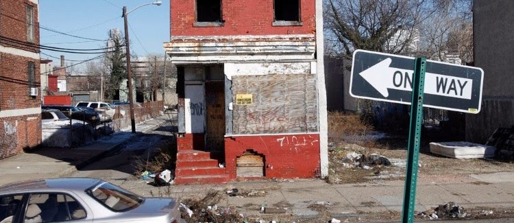 An abandoned, boarded-up red brick building, adorned with graffiti, sits as a testament to the city's attempts to solve poverty. Debris and trash are scattered on the sidewalk. A car is parked nearby, with a one-way street sign in sight. Sparse trees and more buildings loom in the background.