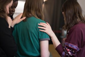 A group of four people stands close together, with two individuals placing their hands supportively on the shoulders of a person in a green shirt, perhaps discussing reasons for drifting away from the church. The setting appears to be indoors, with a soft-focus background.