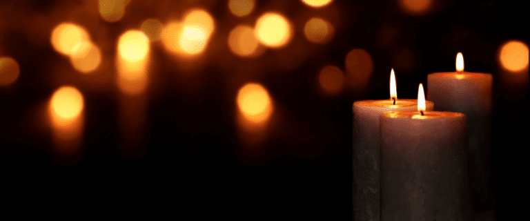 Three lit candles in the foreground emit a warm glow, reminiscent of a Christmas church service, set against a dark backdrop. Many out-of-focus lights create a bokeh effect, adding to the serene and peaceful atmosphere of the scene.
