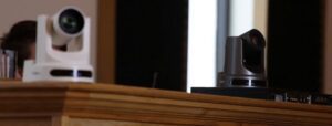 Two PTZ cameras are positioned on a wooden surface, one white and one black, facing slightly toward the left. The background is blurred, with a glimpse of a window or bright area reminiscent of church interiors.