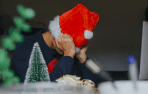 A person sits at a desk, wearing a red Santa hat, symbolizing Christmas stress as they cover their face with both hands. In the foreground, a small Christmas tree decoration and ribbon remind them that holiday tips might just help reduce stress during this festive season.