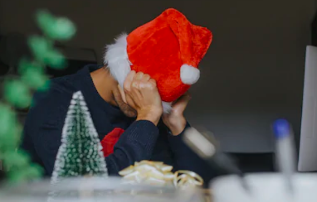 A person sits at a desk, wearing a red Santa hat, symbolizing Christmas stress as they cover their face with both hands. In the foreground, a small Christmas tree decoration and ribbon remind them that holiday tips might just help reduce stress during this festive season.