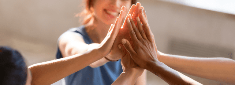 A group of people high-fiving each other, smiling and appearing upbeat. Their hands are the focus in the center, while their faces blur slightly in the background, signifying teamwork and celebrating achieving results together.