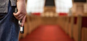 A person holding a handgun stands at the entrance of a church with red carpeted aisles and empty pews, underscoring the urgent need for church safety and preparedness in tense or dramatic situations.