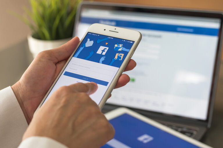 A person holds a smartphone displaying the Facebook login screen, while a laptop in the background mirrors the same page. Beside the laptop, a small plant adds a touch of nature to this digital setting, highlighting how social connectivity bridges our online and offline worlds.