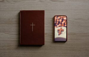 A closed brown book with a cross on its cover rests on a wooden surface next to a smartphone. The phone, often used for live streaming church services, displays an image of a wooden cross placed on an open book with a blurred background.