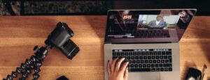A person uses a laptop displaying a live streaming video of a speaker. Next to the laptop, there's a DSLR camera on a flexible tripod. The scene suggests a workspace for video editing or content creation on a wooden desk, likely crafted during the COVID-19 church closure period.
