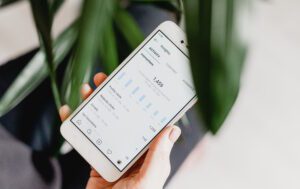 A hand holds a smartphone showcasing an app with analytics, highlighting bar graphs and statistics like impressions and interactions. The display reflects an increase in social media presence. A blurred green plant can be seen in the background.