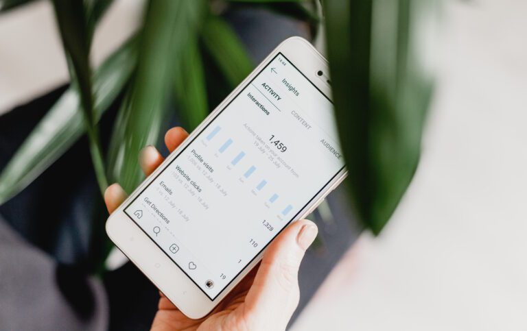 A hand holds a smartphone showcasing an app with analytics, highlighting bar graphs and statistics like impressions and interactions. The display reflects an increase in social media presence. A blurred green plant can be seen in the background.