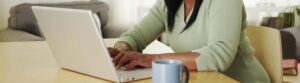 A person wearing a green sweater is typing effectively on a laptop at a wooden table. There's a blue mug in the foreground, and a sofa is visible in the background, possibly ready for some streaming services later.