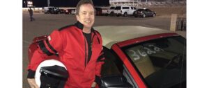 A man in a red and black racing jacket smiles while holding a white helmet. Beside the number 3646 red car, he stands as if ready for action, much like how StreamingChurch.tv is revving up online church services. Several parked vehicles and a fence hint at a racetrack setting.