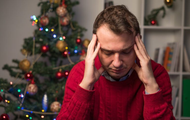 A person in a red sweater sits with their head in their hands, looking stressed, contemplating ways to enjoy Christmas. The decorated tree glows with colorful lights and ornaments, offering a gentle reminder of the festive spirit around them.