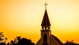 A church steeple with a cross silhouetted against a vibrant orange and yellow summer sunset sky, surrounded by dark trees, inspires hope and renewal, reminiscent of a season of growth.