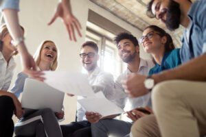 A group of smiling people sitting in a circle indoors, engaging in a lively discussion about training church volunteers. Some are holding papers and a laptop, suggesting a collaborative and positive atmosphere.
