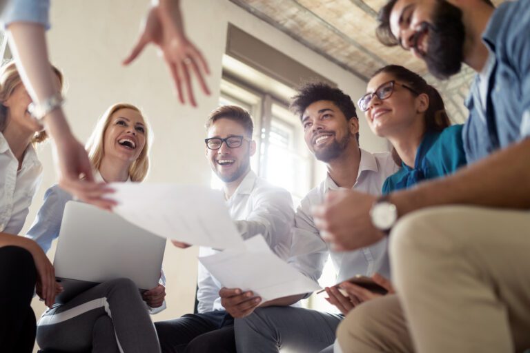A group of smiling people sitting in a circle indoors, engaging in a lively discussion about training church volunteers. Some are holding papers and a laptop, suggesting a collaborative and positive atmosphere.