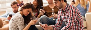 A group of people sits closely together indoors, each focused on their smartphones. Embracing technology, some are lounging on a couch while others sit on the floor. They appear relaxed and engaged with their devices, exemplifying how to reach millennials in today's digital age.