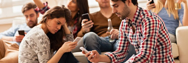 A group of people sits closely together indoors, each focused on their smartphones. Embracing technology, some are lounging on a couch while others sit on the floor. They appear relaxed and engaged with their devices, exemplifying how to reach millennials in today's digital age.