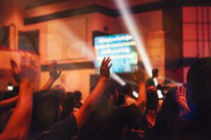 A crowd with raised hands enjoying a live concert in a dimly lit venue, as colorful stage lights dance around, creating a lively atmosphere. It's the perfect backdrop to start streaming and share the electrifying experience with those who couldn't be there.