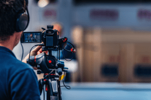 A videographer wearing headphones films with a professional camera on a tripod, perfecting the shot for Church Online streaming services. The background is blurred to emphasize the person’s hands adjusting the camera, while red cables snake around the church streaming setup.