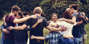 A diverse group of volunteers stands in a circle outdoors, their arms wrapped around each other in a joyful group hug. They bask in appreciation for the sunny day in a park, surrounded by lush green trees.