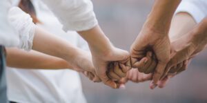 A close-up of a diverse group of people's hands forming a supportive circle. They are joining fists together, symbolizing unity and teamwork, motivating volunteers through shared purpose. The background is softly blurred, emphasizing the hands in the center.