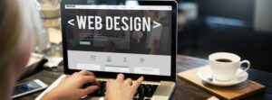 A person is focused on their laptop, which displays a web design page for church websites. A steaming cup of coffee sits nearby on the table.