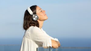 A woman with brown hair leans on a glass railing, eyes closed, wearing white headphones. She's outdoors, enjoying the bright, clear sky and ocean view in the background, dressed in a white shirt—perhaps testing how to enhance church live stream audio with serene surroundings.