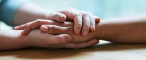 Two women gently clasp hands over a wooden surface, conveying a sense of support and connection in ministry. The image emphasizes empathy and comfort, as they rest their hands together in a caring gesture, embodying women in ministry uniting for strength and encouragement.