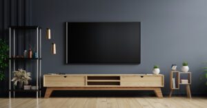 A modern living room features a large flat-screen TV for streaming church services, mounted on a dark gray wall. A wooden media console sits below, adorned with decorative plants and vases. Two pendant lights hang from the ceiling, and a small wooden shelf with an alarm clock stands nearby.