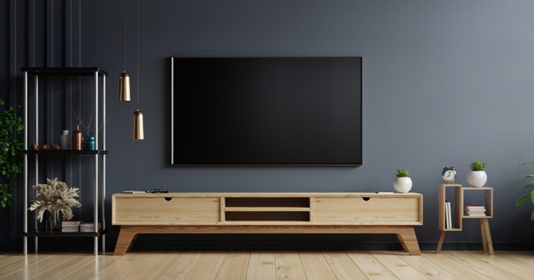 A modern living room features a large flat-screen TV for streaming church services, mounted on a dark gray wall. A wooden media console sits below, adorned with decorative plants and vases. Two pendant lights hang from the ceiling, and a small wooden shelf with an alarm clock stands nearby.