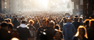 A crowded city street is filled with people walking in various directions, their lively chatter audible beneath the sun’s warm glow. The light creates a silhouette effect on the individuals, while urban structures and a distant church spire line the background.