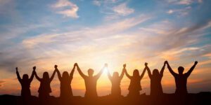 A silhouetted group of church volunteers holds hands and raises them together against a colorful sunset sky, symbolizing unity, togetherness, and shared gratitude.