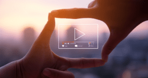 A person's hands form a rectangle against a blurred cityscape at sunset, creating an imaginary screen. A digital overlay shows a play button and video control icons, symbolizing video creation or streaming.