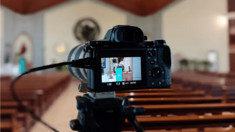 A streaming camera is mounted on a tripod in the church, focusing kindly on the pulpit. The viewfinder captures a speaker at the pulpit, while rows of empty pews stretch into the background.