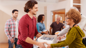 A man in a red shirt hands a box with canned goods to a woman with curly hair in a green sweater, within the lively setting of a church community room bustling with activity, suggesting an atmosphere of great generosity and service.