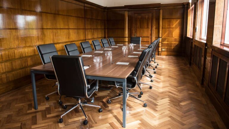 A long wooden conference table with black leather chairs awaits a church board meeting in a room adorned with paneled walls and herringbone parquet flooring. Large windows on the right allow natural light to stream in, highlighting notepads and glasses neatly arranged on the table.