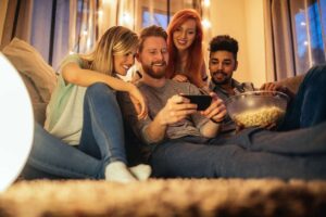 Four friends sitting on a cozy couch, smiling as they enjoy a smartphone video. One holds a large bowl of popcorn, exploring proactive ways to increase online viewership. Warm lighting and string lights create a relaxed and joyful atmosphere.