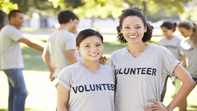 Two happy women wearing grey "VOLUNTEER" t-shirts smile at the camera in a park setting. Other volunteers are blurred in the background, engaged in group activities under the trees, sharing tips and camaraderie.