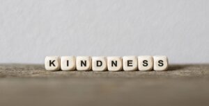 Seven white cubes with black letters spell "KINDNESS" on a wooden surface, highlighting this virtue against a plain light-colored background.
