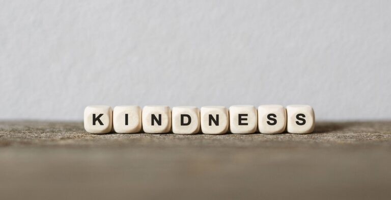 Seven white cubes with black letters spell "KINDNESS" on a wooden surface, highlighting this virtue against a plain light-colored background.