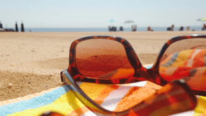 A pair of tortoiseshell sunglasses rests on a colorful striped towel at the beach, embodying classic summertime strategies. The sandy shore and beachgoers under umbrellas stretch out behind them, as the ocean meets the bright blue sky on the horizon.