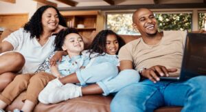 A smiling family of four, with two adults and two children, sits on a couch in a cozy living room. They gather around a laptop as part of their online church community, appearing engaged and happy as they look at the screen.