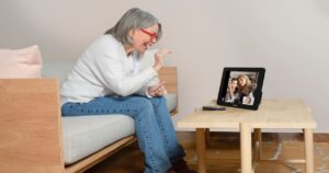 An older woman with gray hair and red glasses is sitting on a sofa, smiling and waving at a tablet. The tablet, resting on a wooden coffee table, displays an engaging video call with three people on the screen—keeping seniors connected and vibrant.
