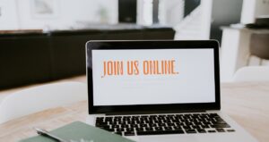 A laptop on a table displays the phrase "Join us online" in orange text, showcasing the small church's commitment to building an engaging online presence. A notebook rests beside the laptop in a modern, well-lit room with sofas and a staircase in the background.