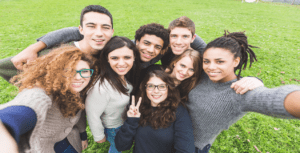 A group of eight young people, diverse in appearance, are smiling while posing for a selfie outdoors in 2023. They stand close together on a grassy church lawn, looking happy and relaxed. The person in front is making a peace sign with their hand.