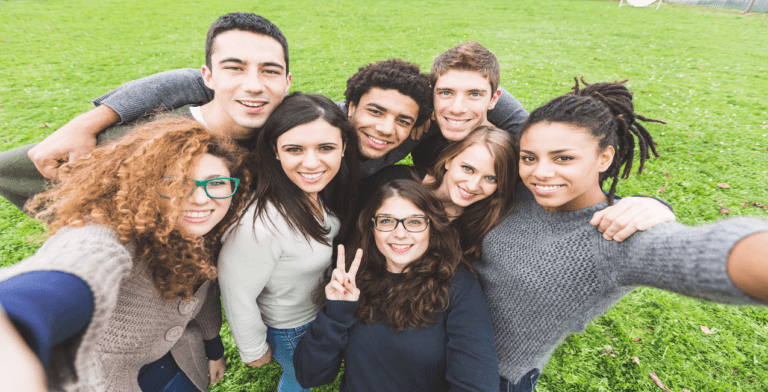 A group of eight young people, diverse in appearance, are smiling while posing for a selfie outdoors in 2023. They stand close together on a grassy church lawn, looking happy and relaxed. The person in front is making a peace sign with their hand.