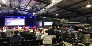 A video camera captures a church service in a modern auditorium with multi-camera video production. A screen displays "Tithes & Offerings," as people stand and listen beneath industrial-style lighting and visible ceiling beams.
